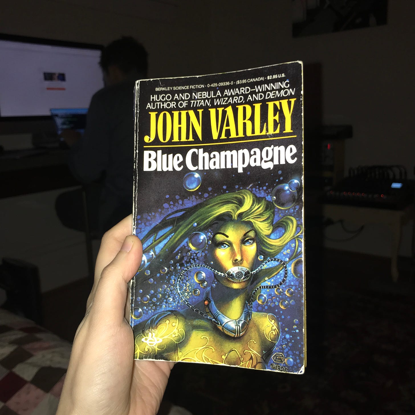My hand holding a worn out copy of John Varley's short story paperback "Blue Champagne."