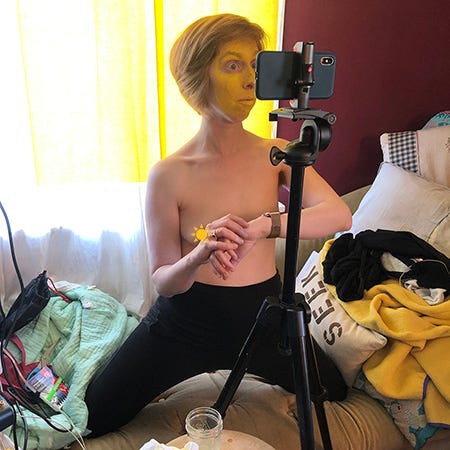 still photo of ilyse on a couch taking pictures of her face painted yellow from an iPhone mounted on a tripod. 