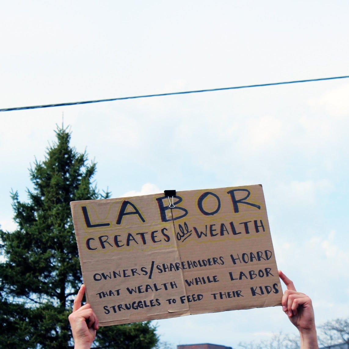 hands holding up a cardboard sign with text reading "LABOR creates all wealth, owners/shareholders hoard that wealth while labor struggles to feed their kids"
