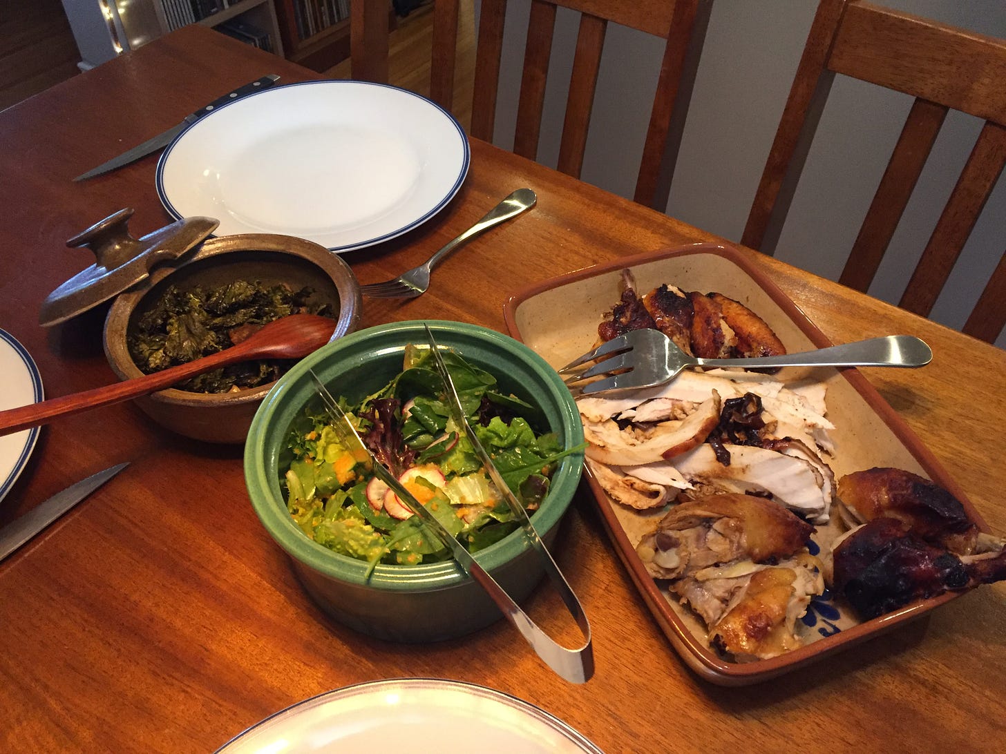 A table set for dinner with three serving dishes in the middle: one small tureen with crispy kale and mushrooms, a green ceramic bowl of salad, and a rectangular dish with carved pieces of chicken.