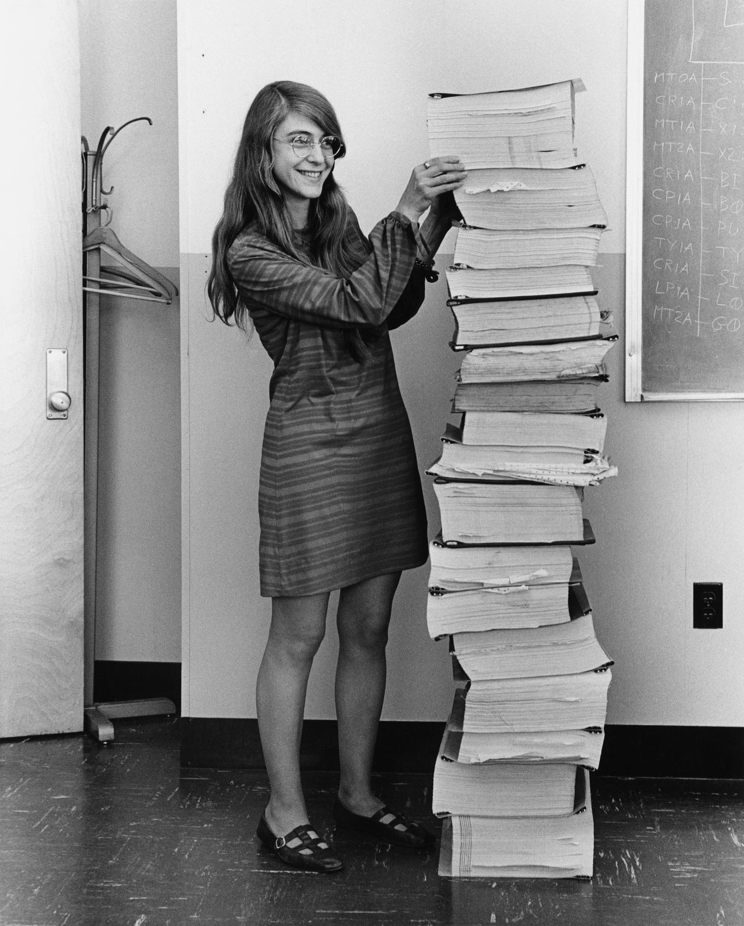 Margaret Hamilton standing next to the navigation software that she and her MIT team produced for the Apollo Project, which is an enormous stack of pages going from the floor up to her head.