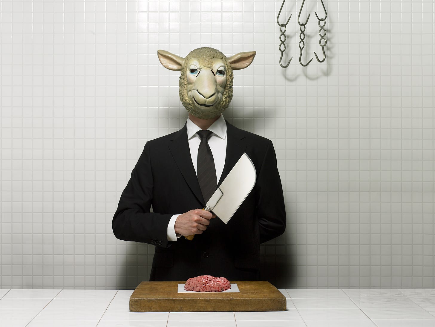 person in black suit and lamb mask holding a cleaver over a small butcher block with ground meat on it
