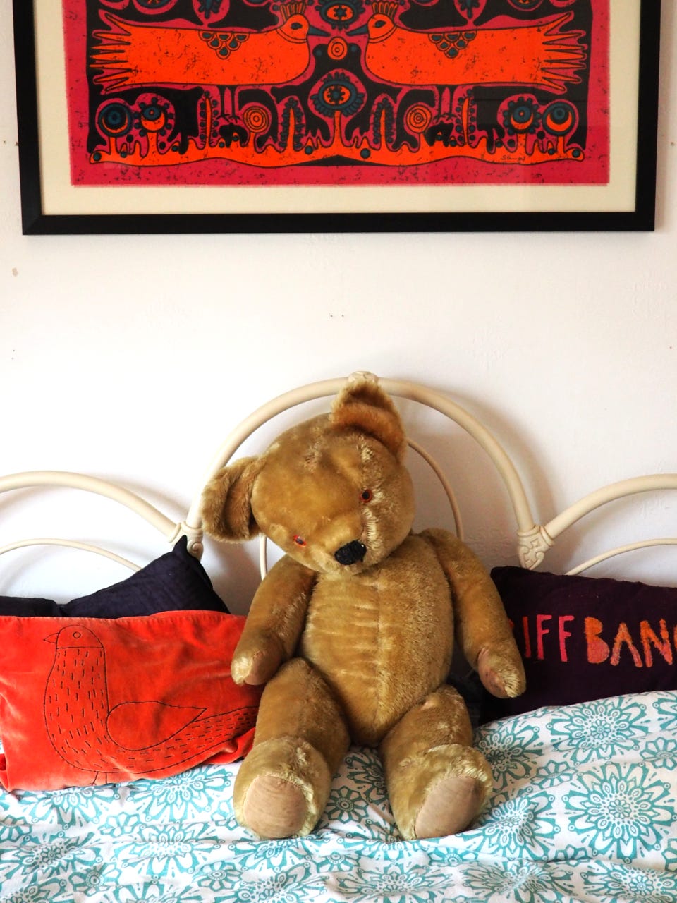Merrythought bear sitting on a bed in front of a white wall with a fraed picture of two orange birds hanging above .