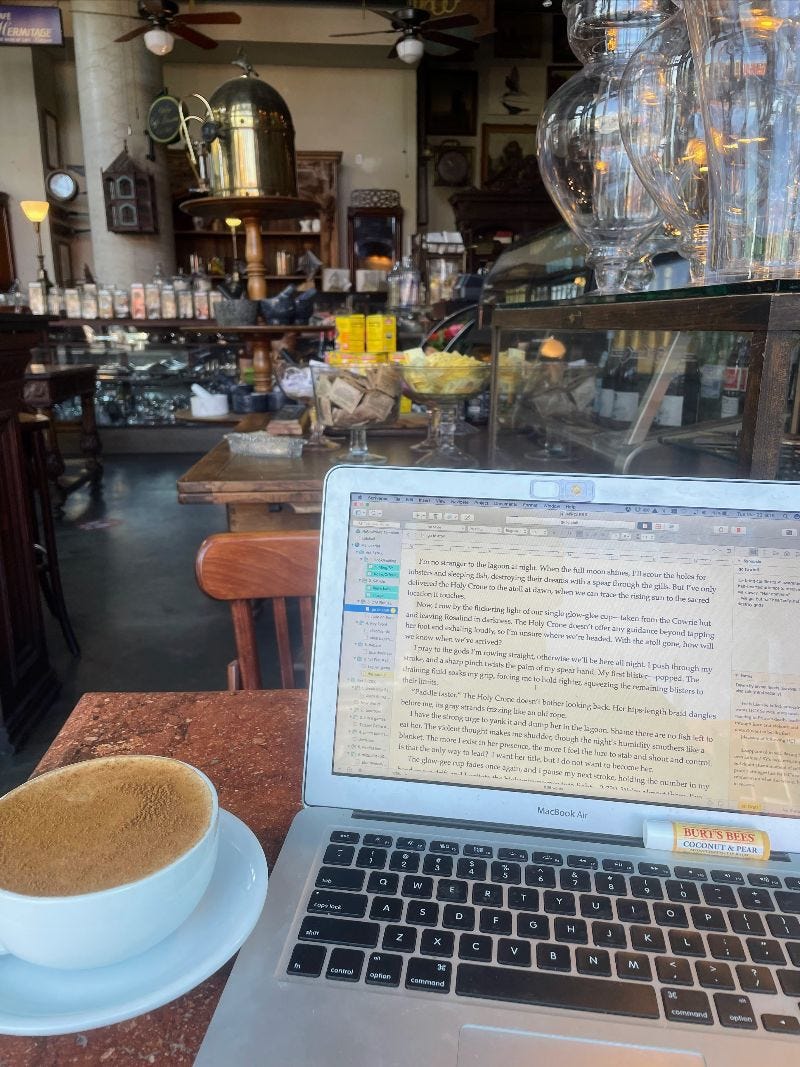 a laptop on a cafe table, a manuscript on the screen. Chai tea latte on the table, and in the background, a cafe with glass jars of loose leaf tea