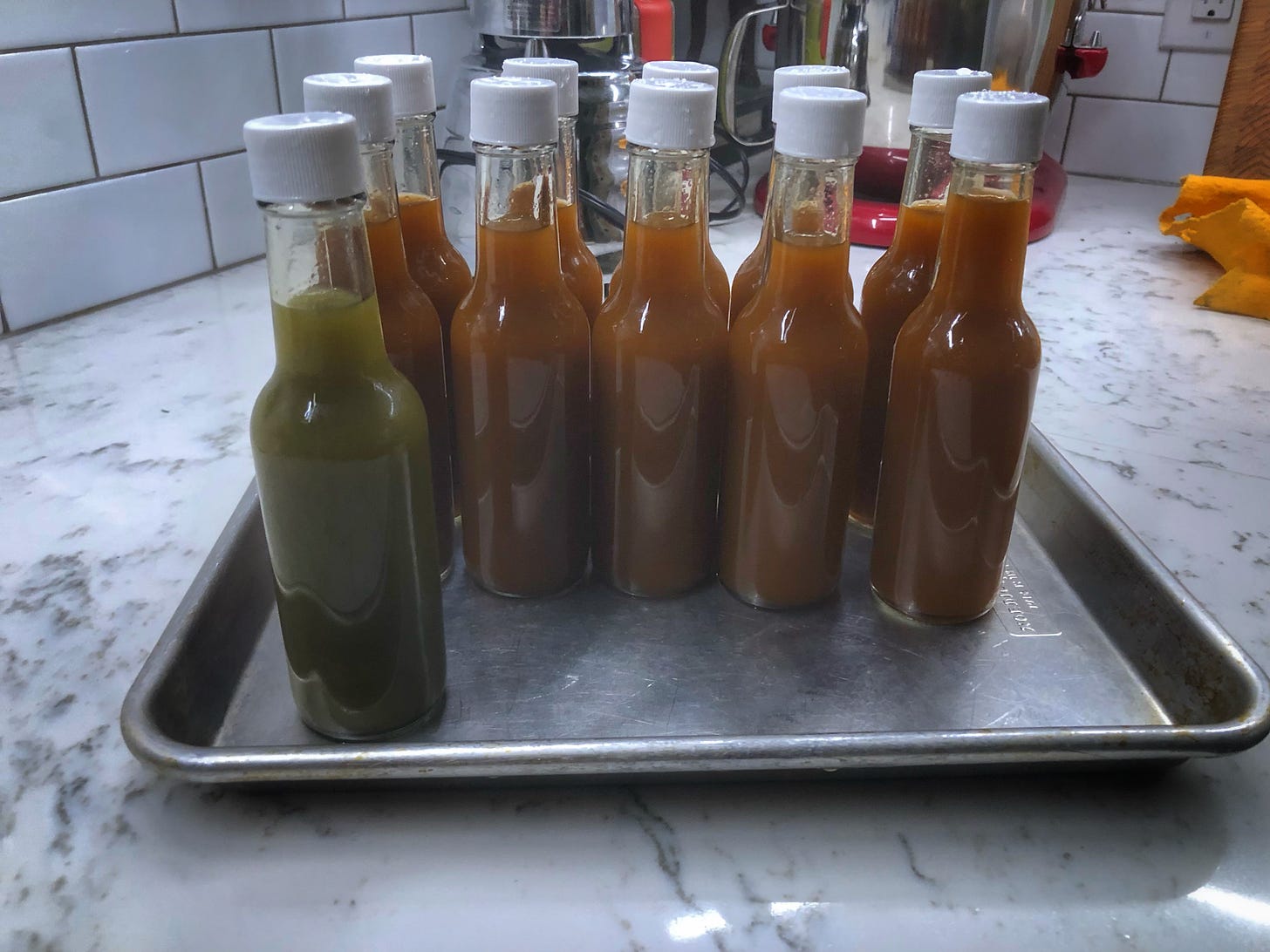 Bottles of hot sauce on a metal tray. The sauce in one bottle is green while the others are russet.