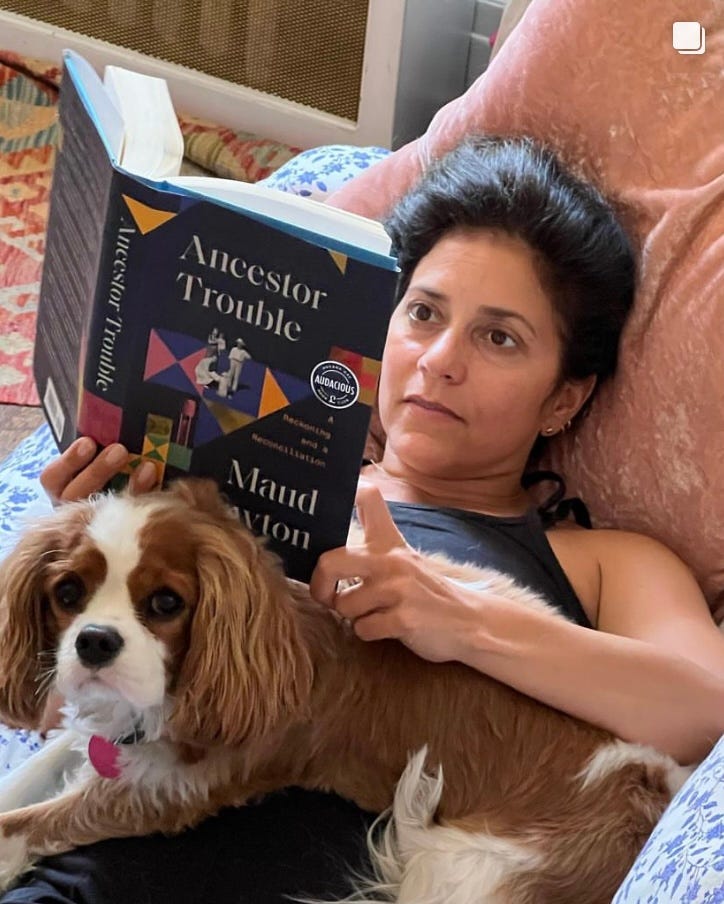 A white woman with dark hair reads Ancestor Trouble while lying on a sofa with a dog on her chest.