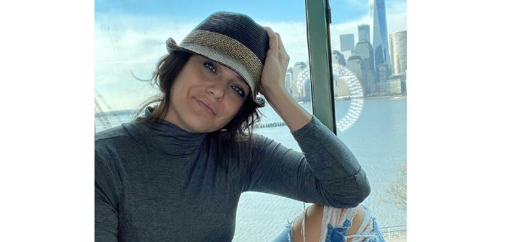 A woman in a hat, with her arm bent, elbow on her knee, and hand on her head, smiling in front of a window with a cityscape and water in the background