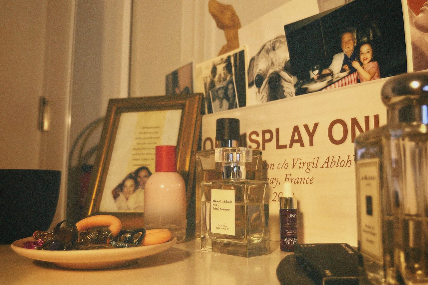 The author's dresser scatered with pefume, a jewlery catchall, and image of her and her mother, and a champagne box filled with books and postcards.