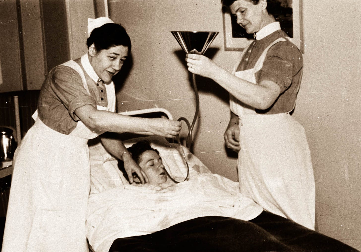 Insulin being administered by two nurses in uniforms. One is holding a funnel and the other is directing the tube attached to the funnel into the patient's nose. Like a beer bong, kind of. 