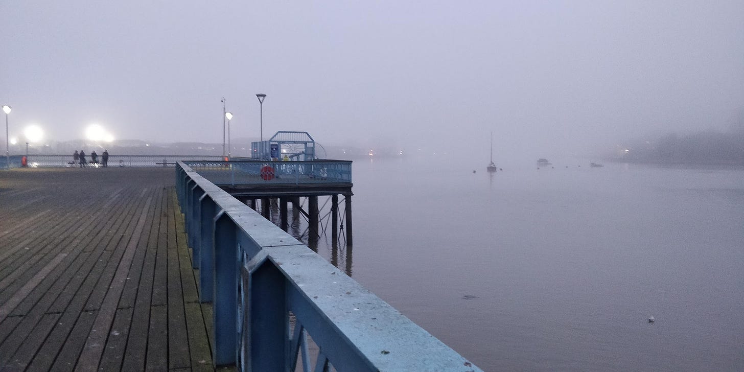 A blue pier. The boards are made of old, algae-ridden wood, and the sides of the pier have started to rust. It is foggy in the background, and lights flare from the distance. It is altogether dreary.