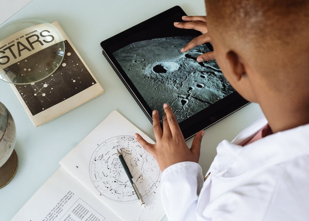 over the shoulder shot of someone looking at a picture of the moon on a tablet, with astronomy books and notebooks in front of them on a white desk