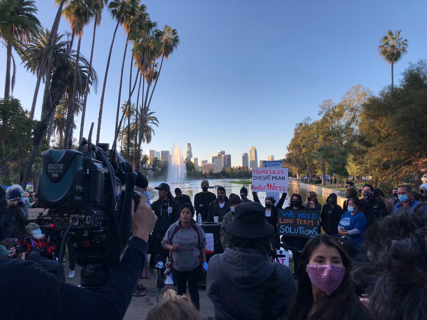 News cameras videotape a crowd of people holding a press conference. Palm trees and skyscrapers in the background.
