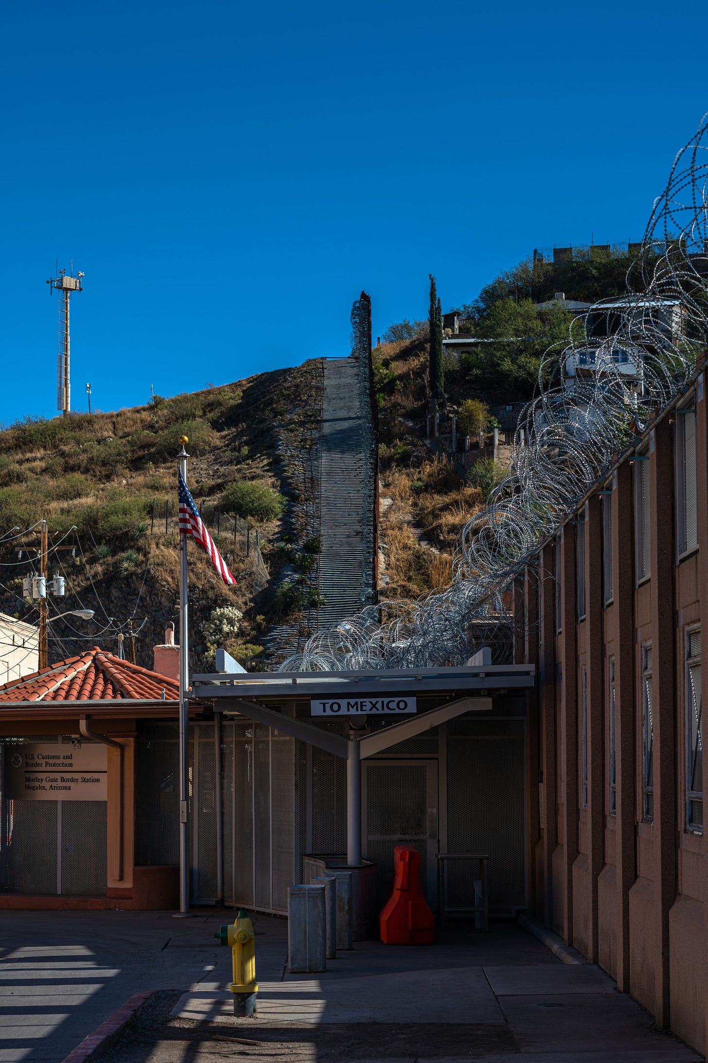 A picture looking along the US/Mexico border wall, as it runs up and over a hill silhouetted against a perfectly clear blue sky. The fence is topped with coils of razor wire and a tall surveillance tower is visible in the near background. A picture looking along the US/Mexico border wall, as it runs up and over a hill silhouetted against a perfectly clear blue sky. The fence is topped with coils of razor wire and a tall surveillance tower is visible in the near background.