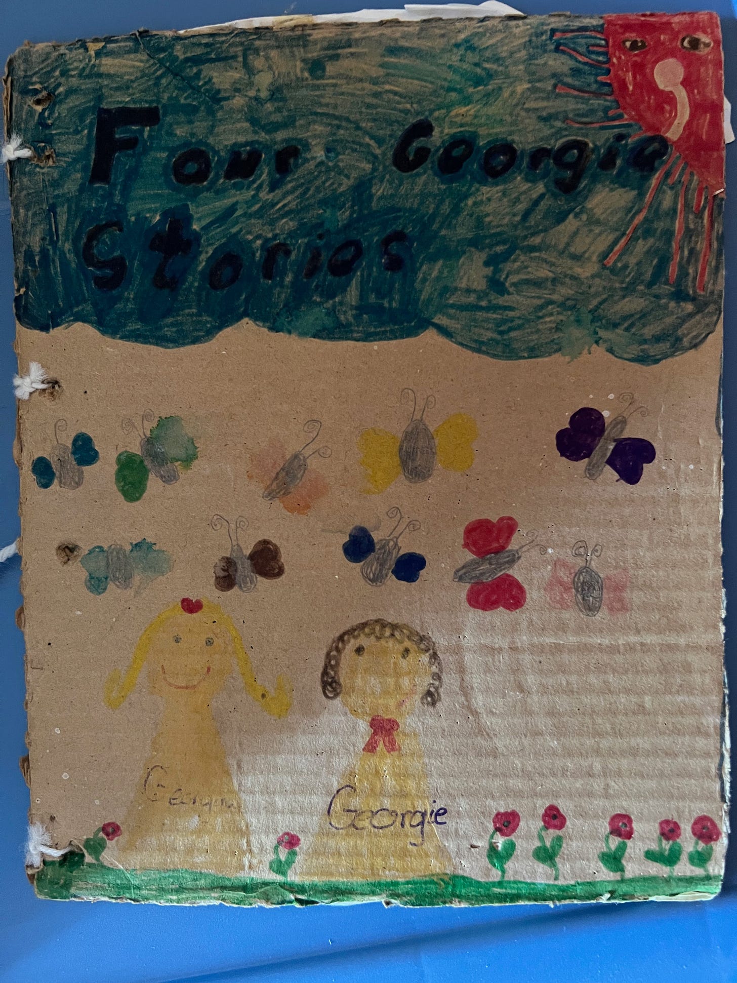 Cover of a "book" bound together with white yarn: a cardboard cover with "Four Georgie Stories" written on a marker drawing of a cloud above several butterflies and two ghosts with the names Georgie and Georgina written on them, some kind of red flowers below