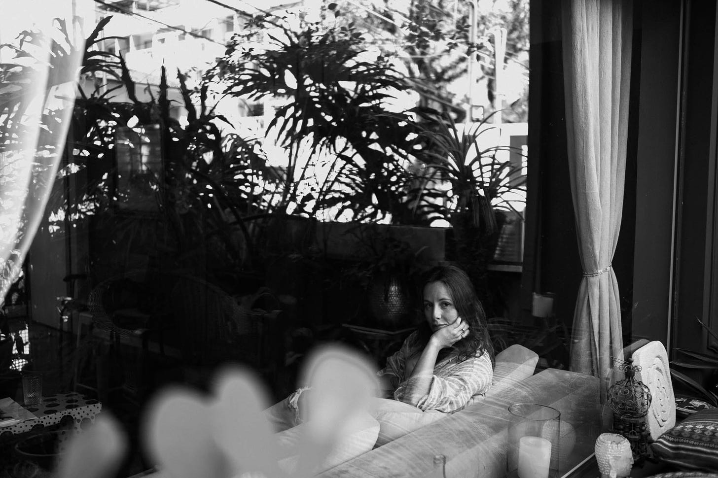 Mulher sentada em sofá atrás de vidraça olhando pela janela através de plantas, imagem em preto e branco