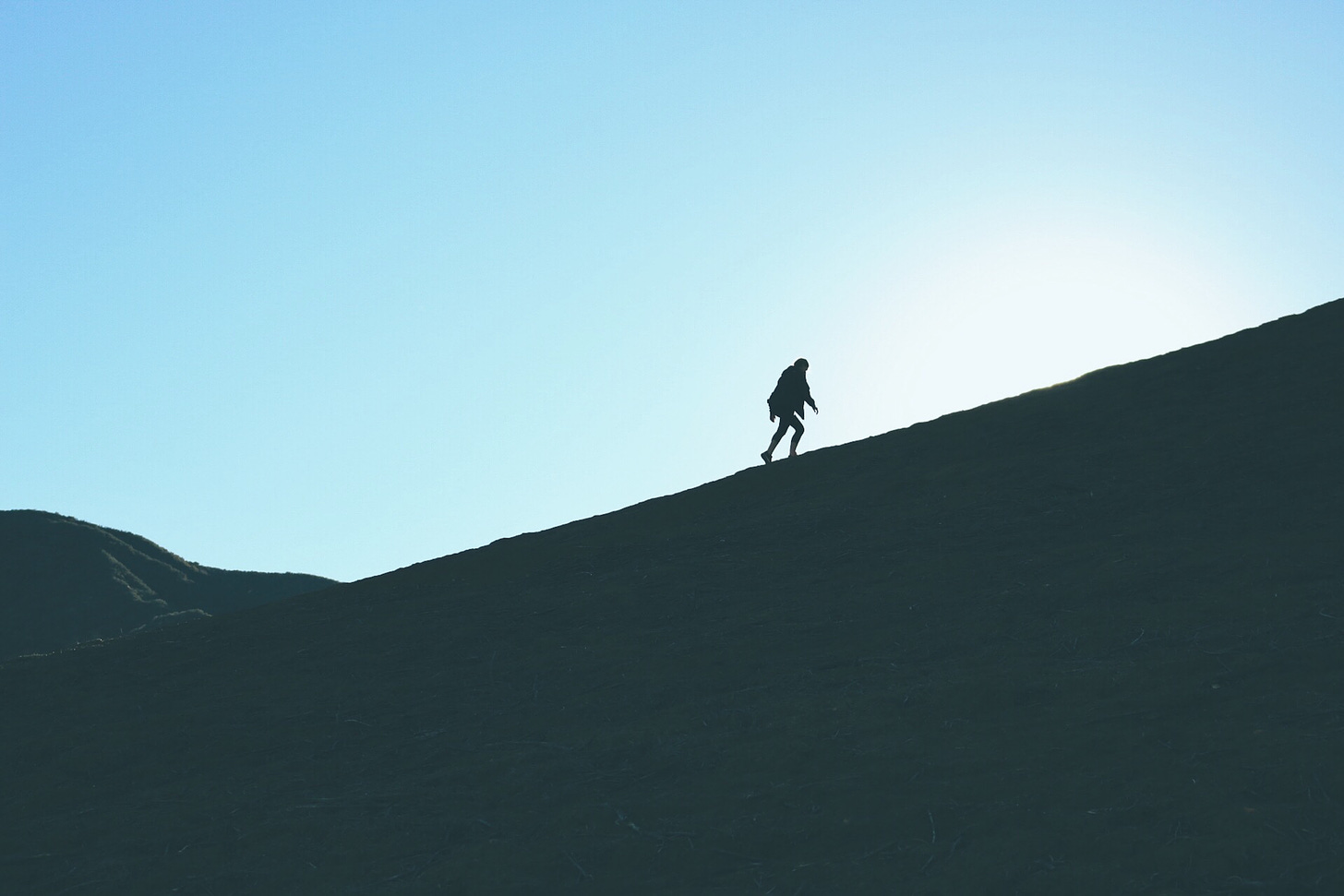 A person, shown from the side, climbs a mountain. The sky is bright behind them so they're shown in silhouette.