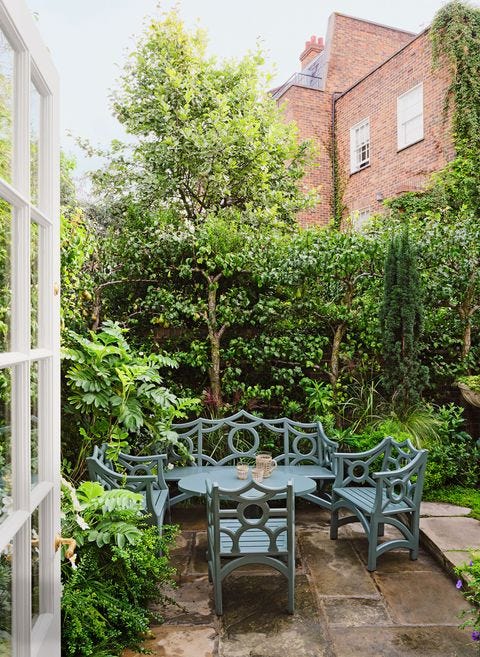 the garden is protected by a wall of small trees and shrubs, on the stone flooring sits a round table surrounded by three chairs and a bench with open decorative backs painted in french blue