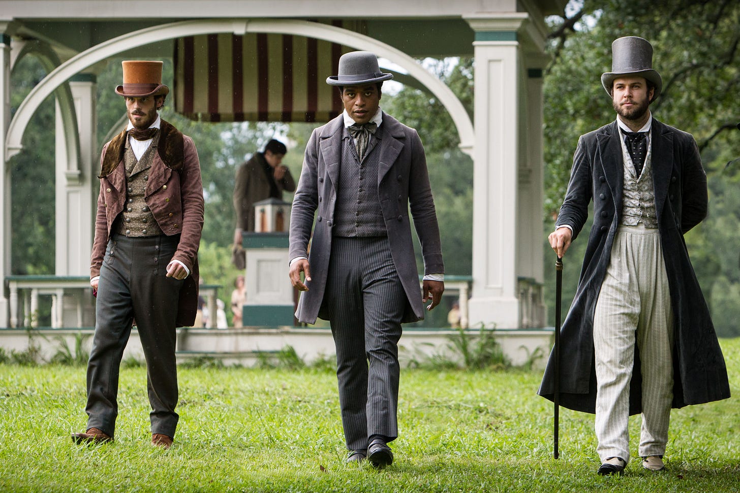 This image shows Merrill Brown (played by Scoot McNairy) on the left, Solomon Northup (played by Chiwetel Ejiofor) in the center, and Abram Hamilton (played by Taram Killam) on the right. All three men are dressed in period finery (circa 1841) and walk through a public park in Saratoga Springs, New York. This image shows Merrill Brown (played by Scoot McNairy) on the left, Solomon Northup (played by Chiwetel Ejiofor) in the center, and Abram Hamilton (played by Taram Killam) on the right. All three men are dressed in period finery (circa 1841) and walk through a public park in Saratoga Springs, New York.