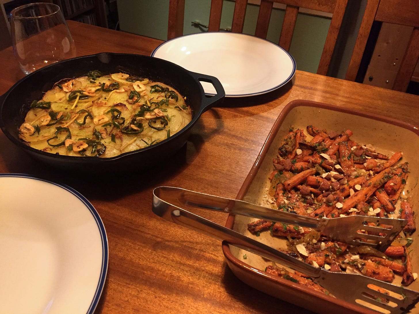 a cast iron pan of potato gratin topped with fried chilies and garlic sits between two empty plates. To the right, a serving dish of carrots speckled with herbs and chopped nuts.