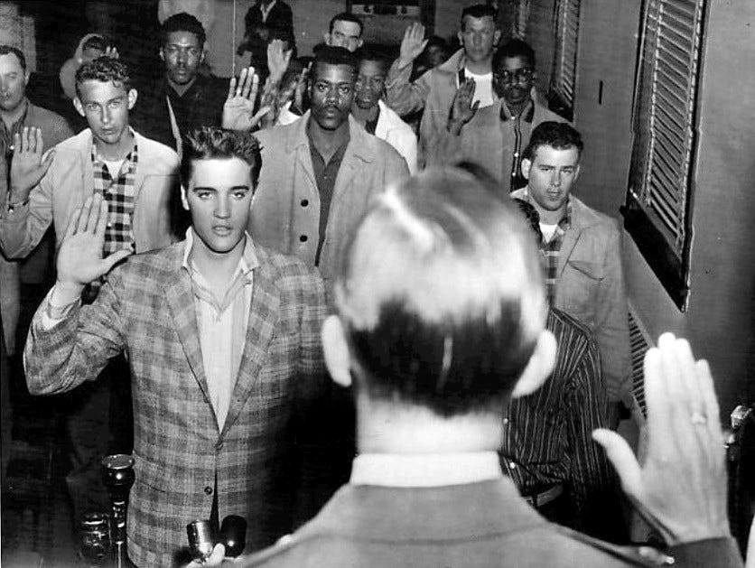 Elvis is sworn into the army, along with others.  The group of men stand with their right hands up in the air as they take their oath of enlistment.