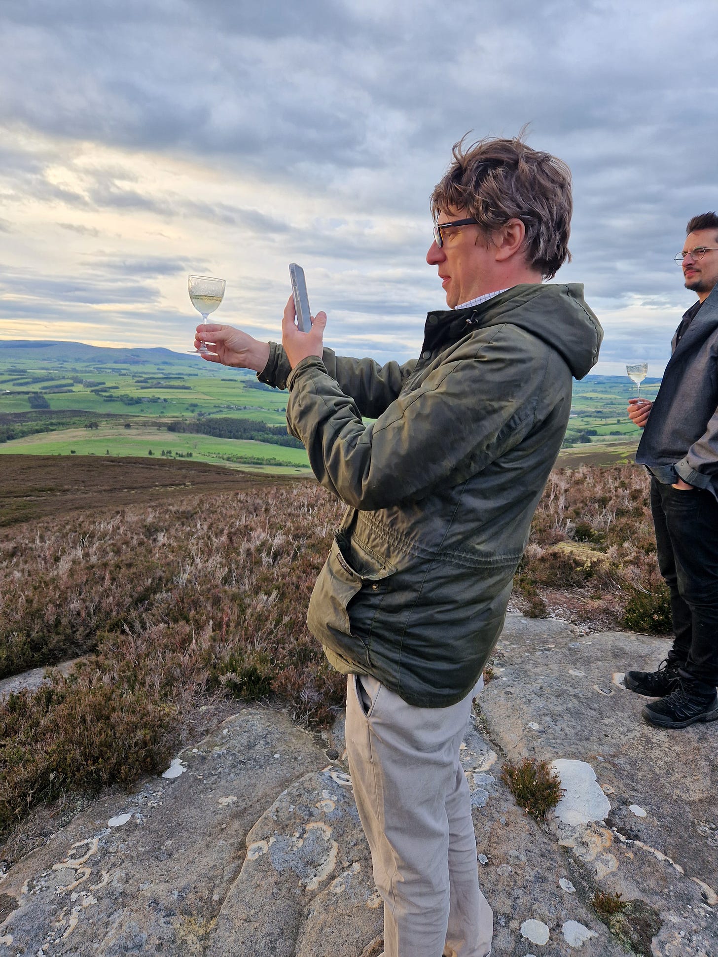 Chris Garden, Head Distiller at Hepple, stands on a hilltop above an expansive moorland vista. He is holding a martini aloft and is photographing it with his phone.