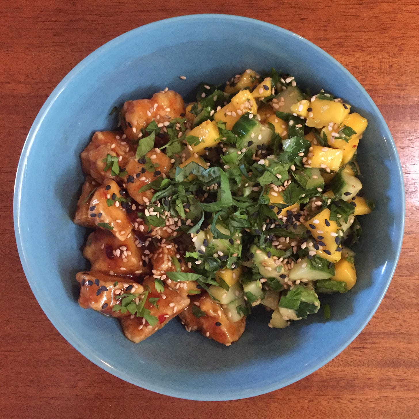 A blue bowl with pieces of crispy tofu in sweet chili sauce on one half, and a mango-cucumber salad with herbs and avocado on the other half. Basil and cilantro and 2 colours of sesame seeds are scattered across the top.