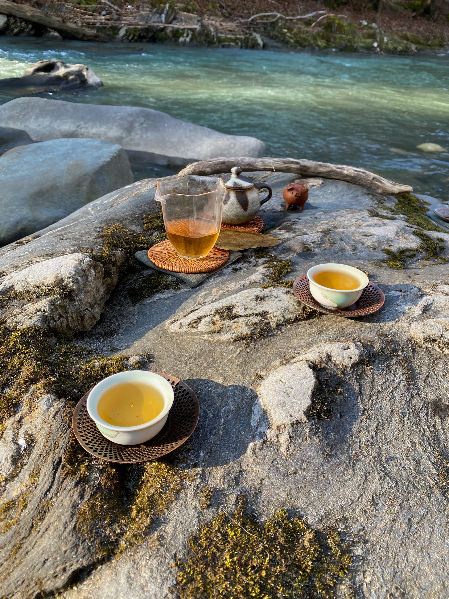 A selection of teaware sits on a large flat rock at the edge of a rushing river: two small ceramic cups filled with golden tea, a glass pitcher filled with the same tea, a small ceramic teapot, and a brown pig figurine. 