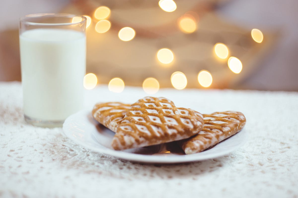 Two heart-shaped gingerbread biscuits on a plate on a white embroidered tablecloth beside a glass of milk, with a Christmas tree in the background.