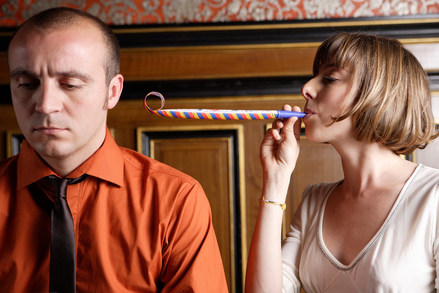 A short haired woman blowing a party noisemaker at the ear of a decidedly unamused man in a tie.