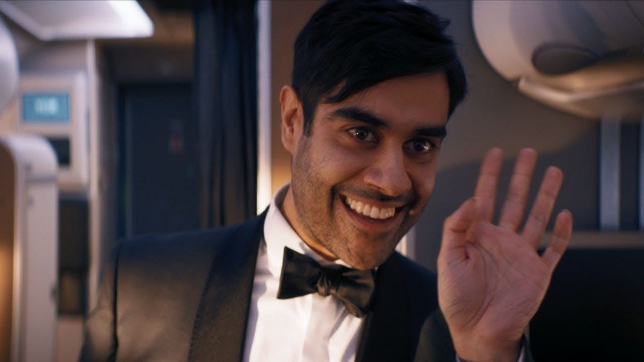 This image shows Sacha Dhawan as the character "O," dressed in a tuxedo and waving hello at the offscreen Doctor, while standing in the central aisle of a jetliner owned by Daniel Barton's VOR Corporation.. This image shows Sacha Dhawan as the character "O," dressed in a tuxedo and waving hello at the offscreen Doctor, while standing in the central aisle of a jetliner owned by Daniel Barton's VOR Corporation..
