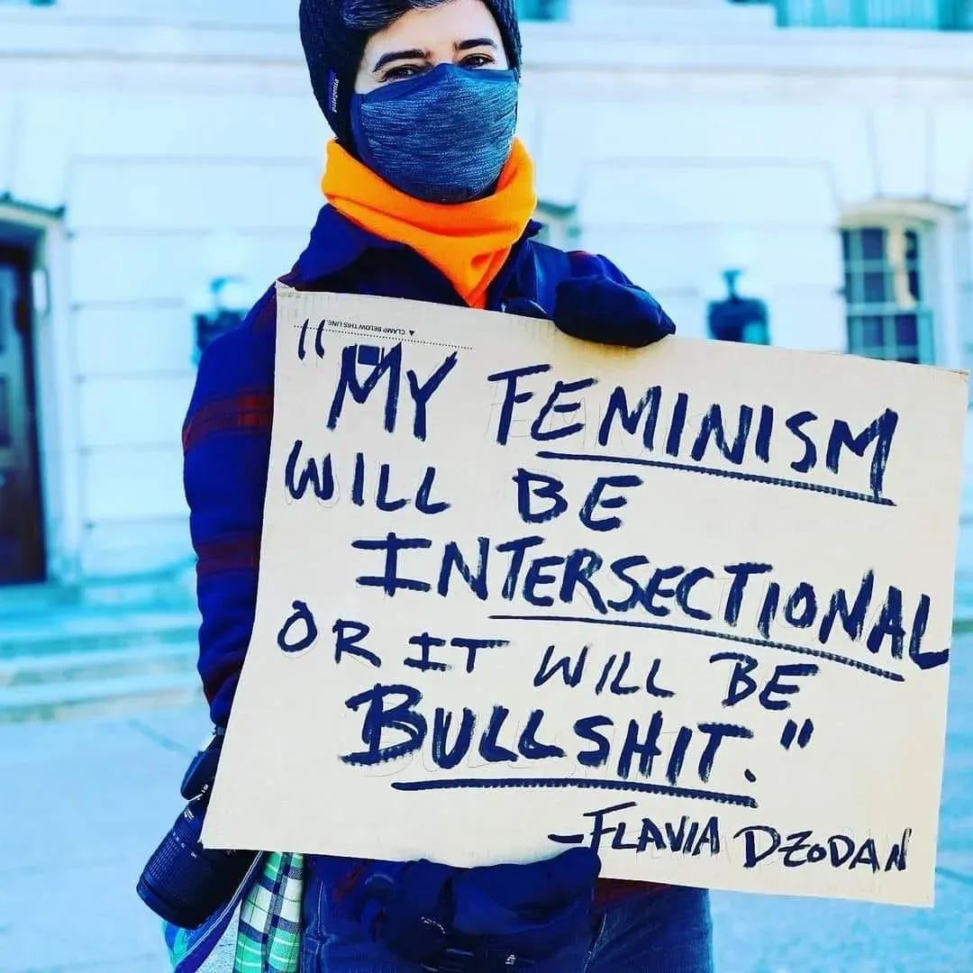 A bundled up and masked Emily stands holding a sign that reads "My feminism will be intersectional or it will be bullshit - Flavia Dzodan"