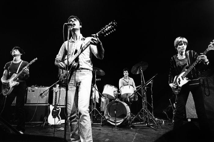 talking heads performing in their early years: front centre, david byrne in pale shirt and trousers playing guitar; back centre, chris frantz on drums; left, jerry harrison in dark clothing playing guitar; right, tina weymouth in dark clothing and with short blonde hair playing bass