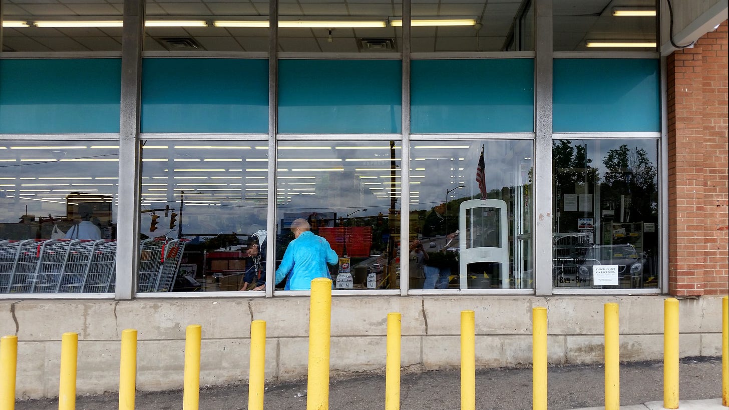 yellow traffic beams in front of tall windows of a grocery store entrance nearby a line of carts and a woman with a bright blue shirt