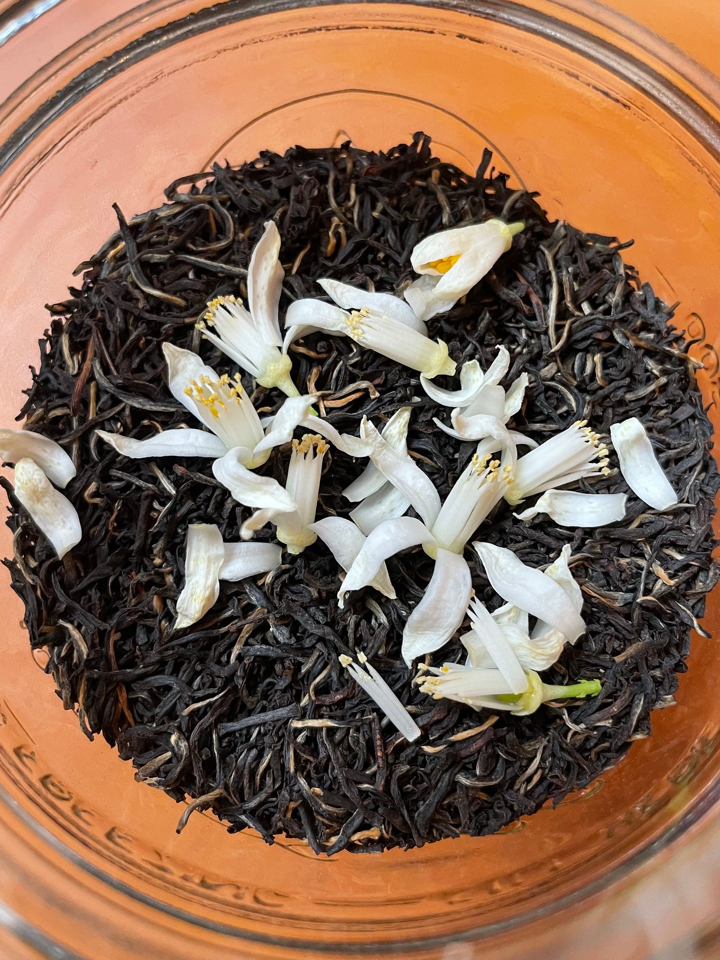 ID: Overhead photo of loose tea leaves and white citrus blossoms in a jar