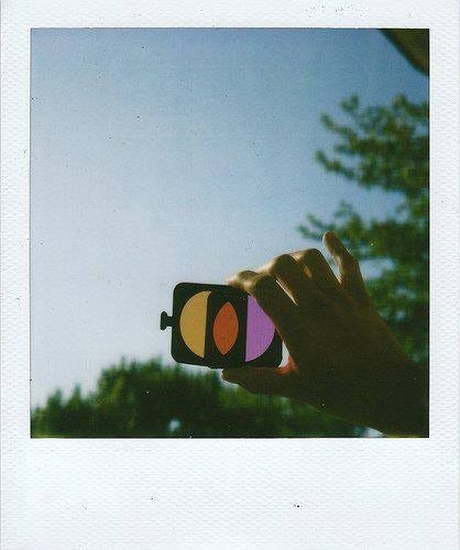A Polaroid of a hand holding two colored filters, one yellow and one magenta, combining to create a third red filter, against the backdrop of a blue sky.
