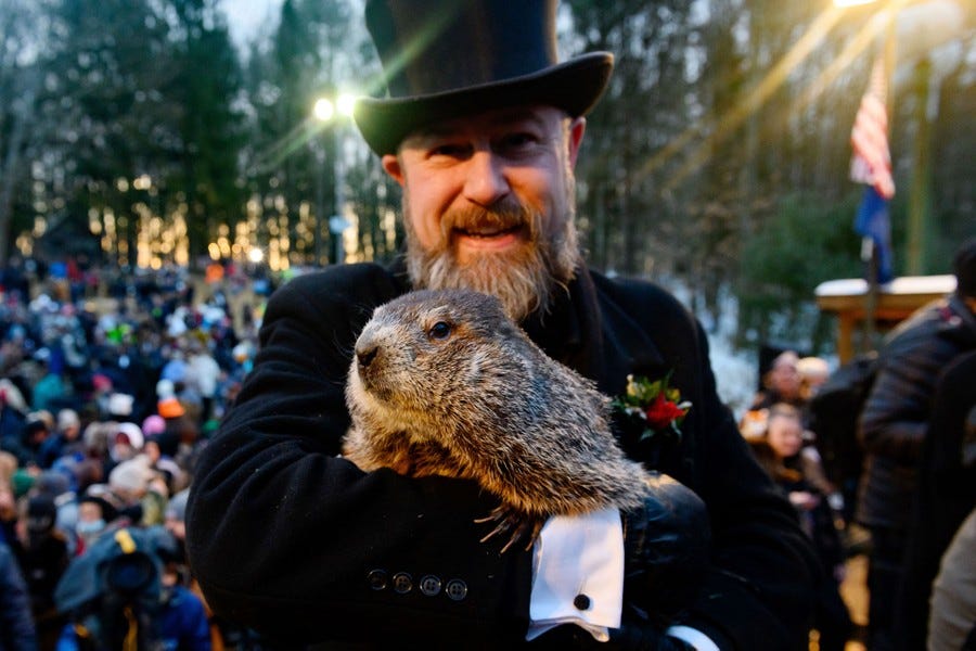 A man wearing a top hat holds a groundhog in his arms, with a large crowd seated behind them.