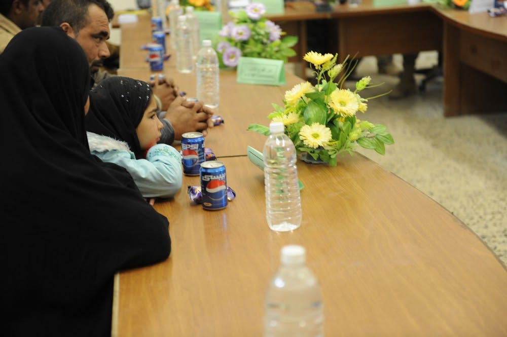 A child, together with her mother, sit at a table. There are flowers, water bottles, cans of Pepsi, and individually wrapped snacks on the table. The mother and daughter are there to recieve condolence pay.