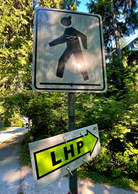 a photo of a road sign with a pedestrian crosswalk warning sign. Below it is a neon sign pointing down a path. The sign says “LHP” on it. 