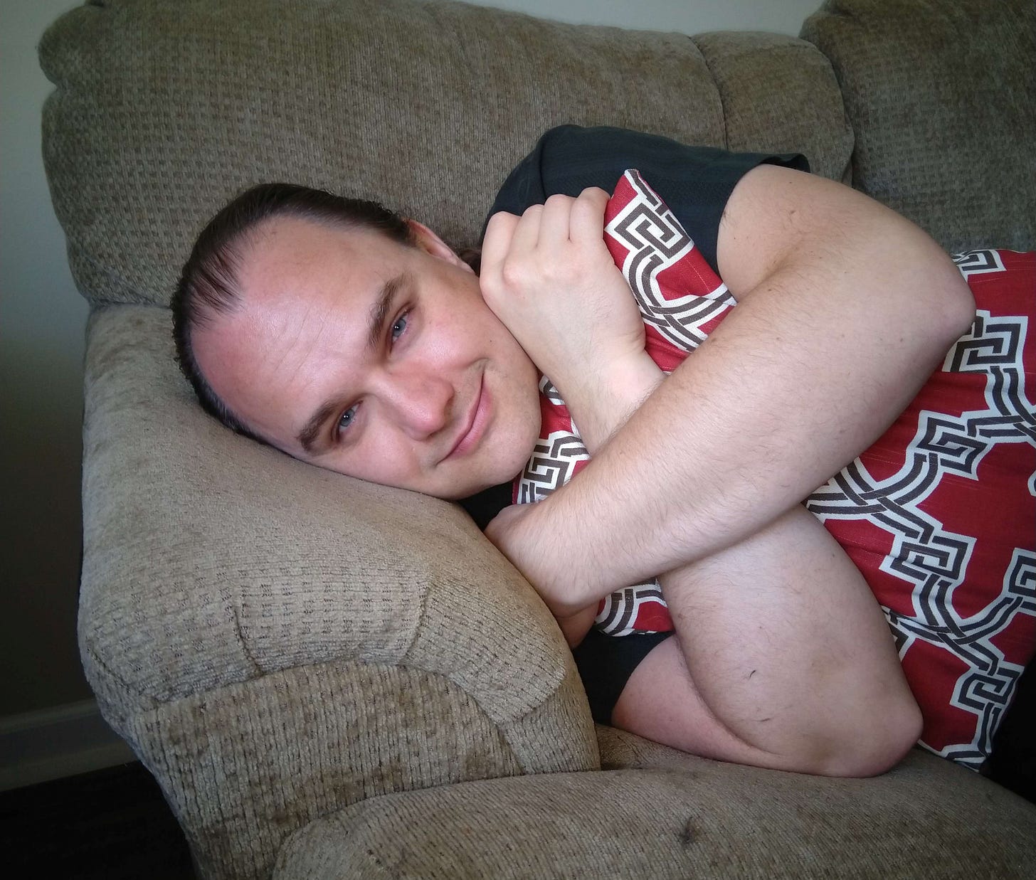 A picture of the author, smiling fully, lying on a loveseat and clutching a pillow with arms crossed.