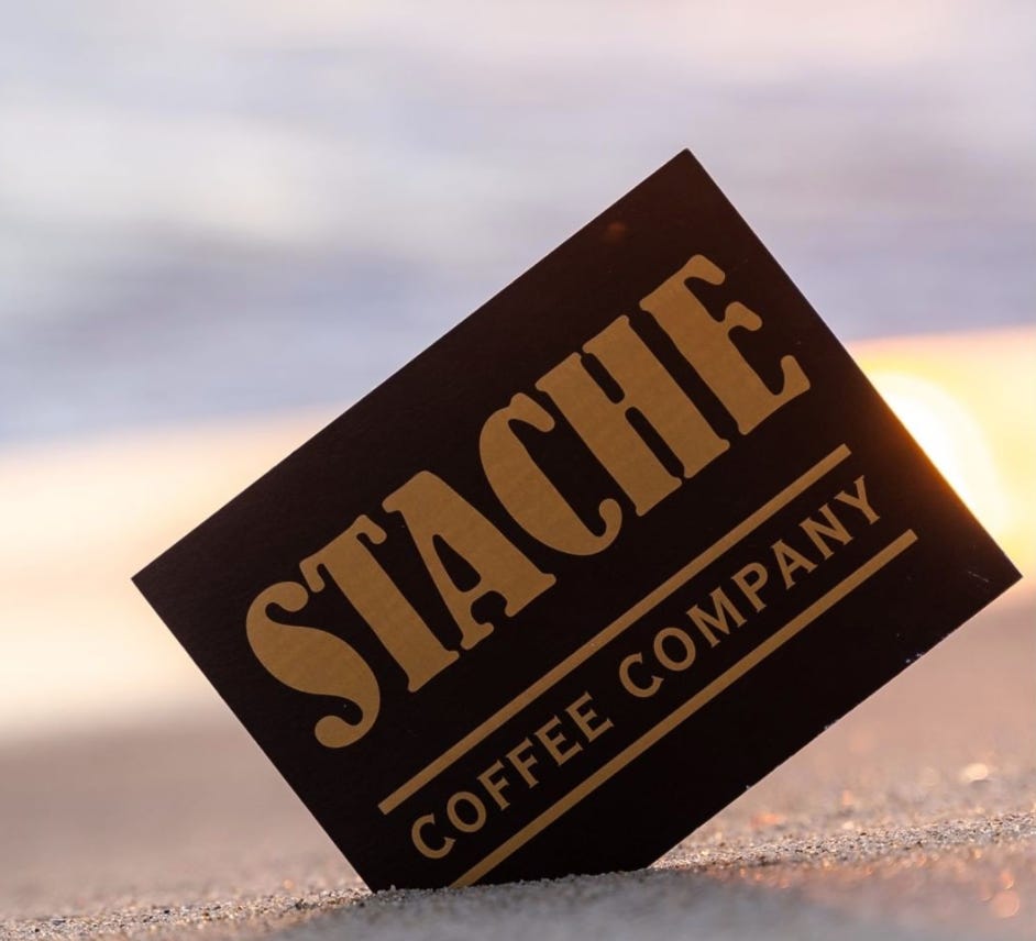 Stache Coffee Company logo sticker stuck in the sand at the beach in front of the sunset.