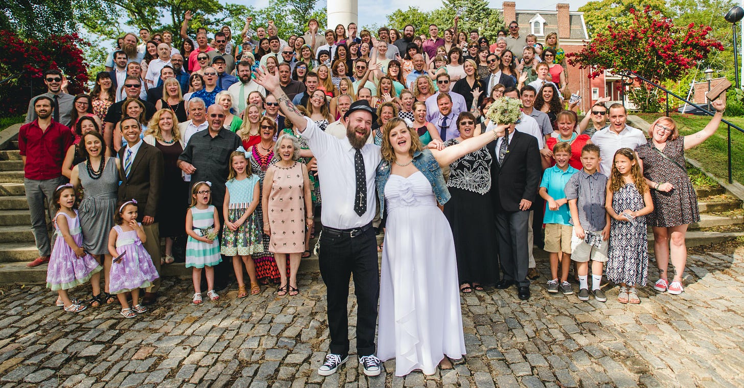 Photo of Deanna and Matt's huge wedding party, standing on stairs behind the couple in their punk rock wedding attire. They have arms outstretched and sooo much joy. Getting this photo is just like what you need to do in your business: try. 