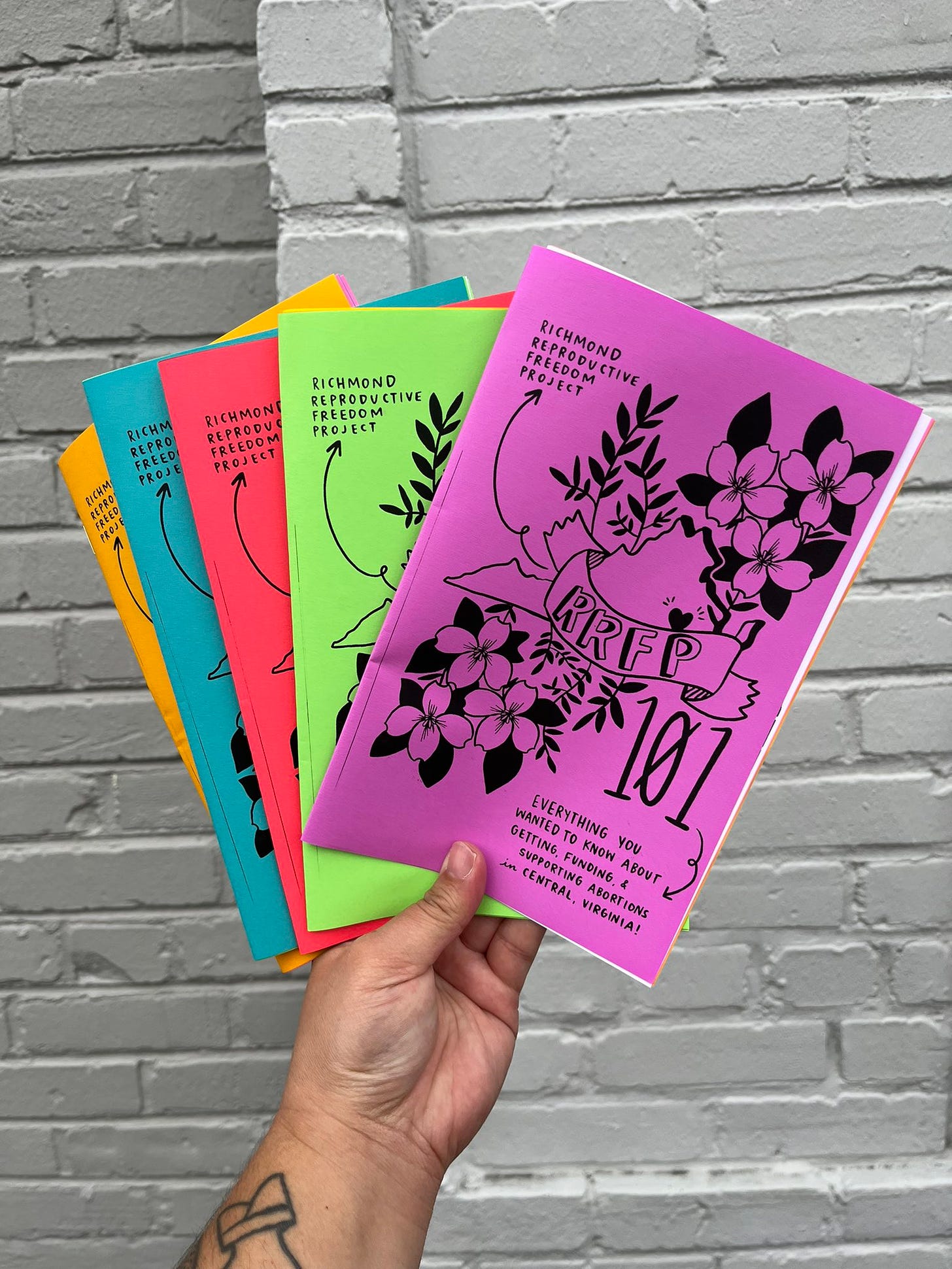 Photograph of a hand holding multiple copies of brightly colored "RRFP 101" zines
