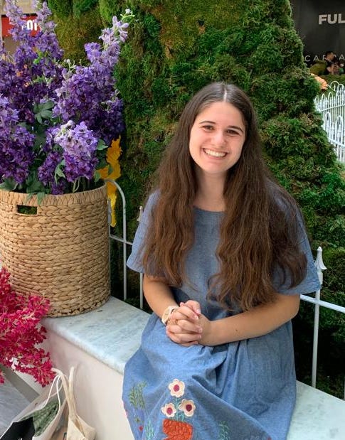 Tari Sztokman, young woman with broad smile and long brown hair falling from her shoulders, sitting against a beautiful backdrop of colorful flowers