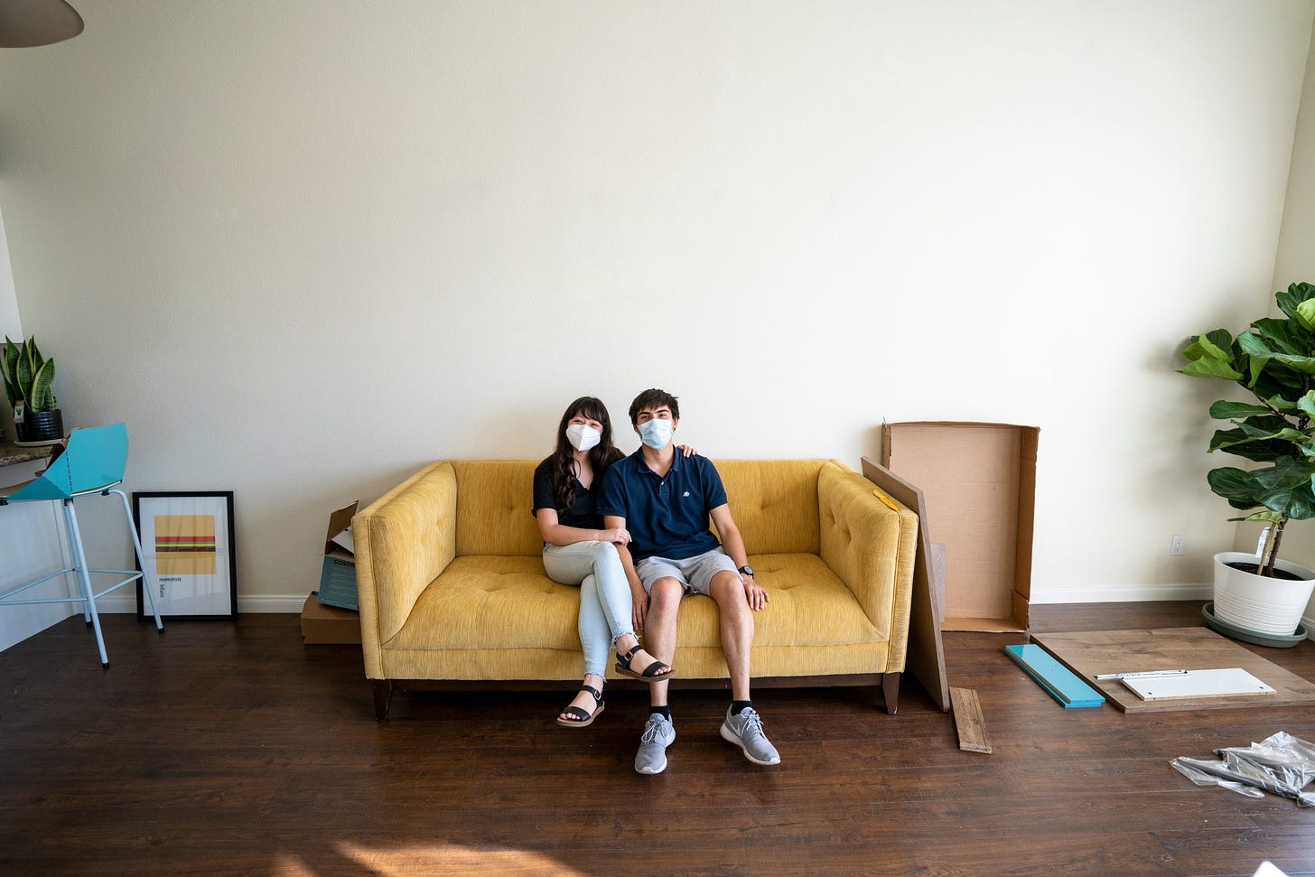 A couple in masks sit on a couch in the middle of a cluttered living room.