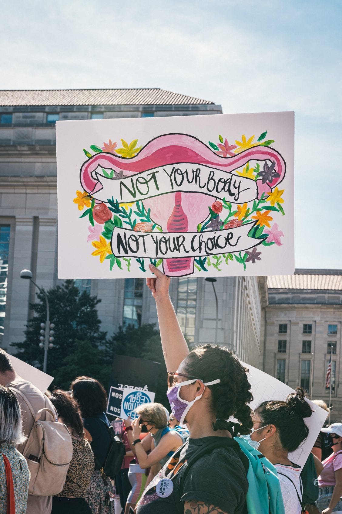 Focus on protest sign with painted uterus and fallopian tubes, across which is written in 2 separate banners " Not your body" "not your choice". Sign held high above head of women marching in US city.