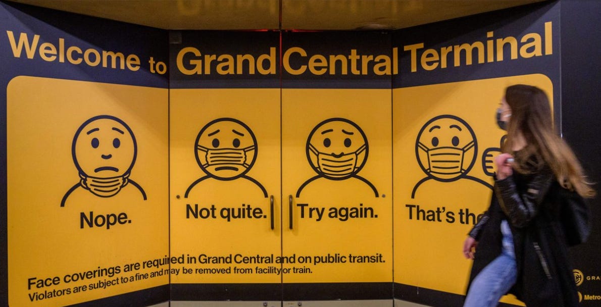 A sign that says Welcome to Grand Central Terminal, face coverings are required in grand central and on public transit. Violators are subject to a fine and may be removed from facility or train The picture is of cartoon figures one wearing mask on chin under it says Nope another figure has the mask on but not covering mouth under it says Not quite Another figure has the mask under the nose and under that it says Try again and the last figure has the mask on properly over nose and mouth and under it says Thats the and the rest of the text is obscured by a person walking past the sign in the photo