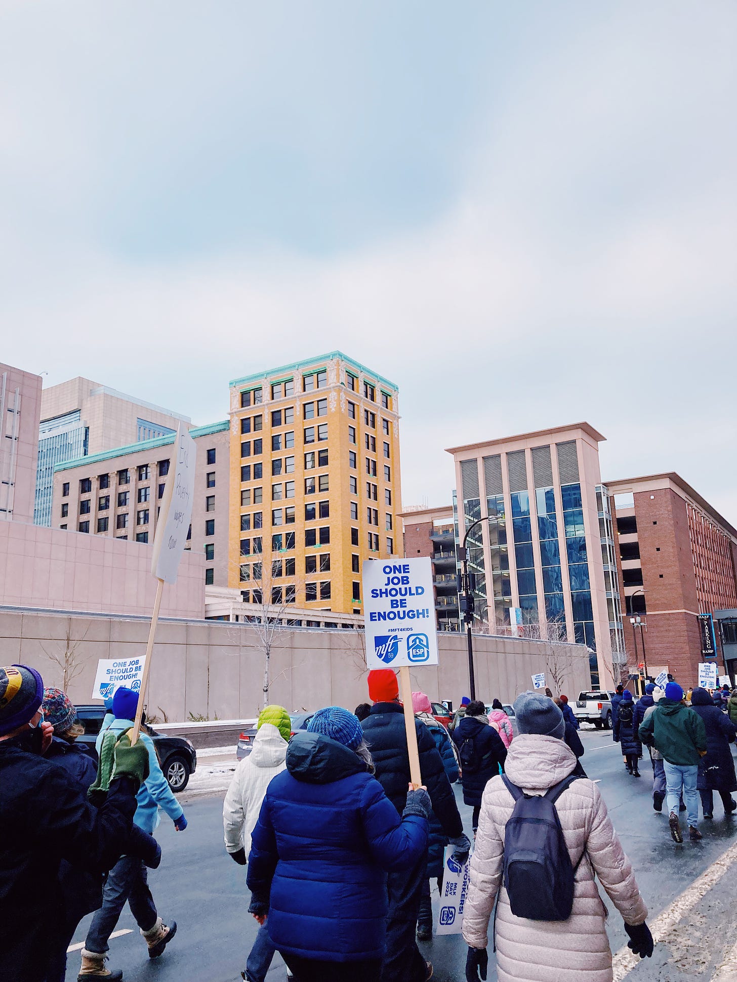 protestors walk in downtown, one sign says "one job should be enough"