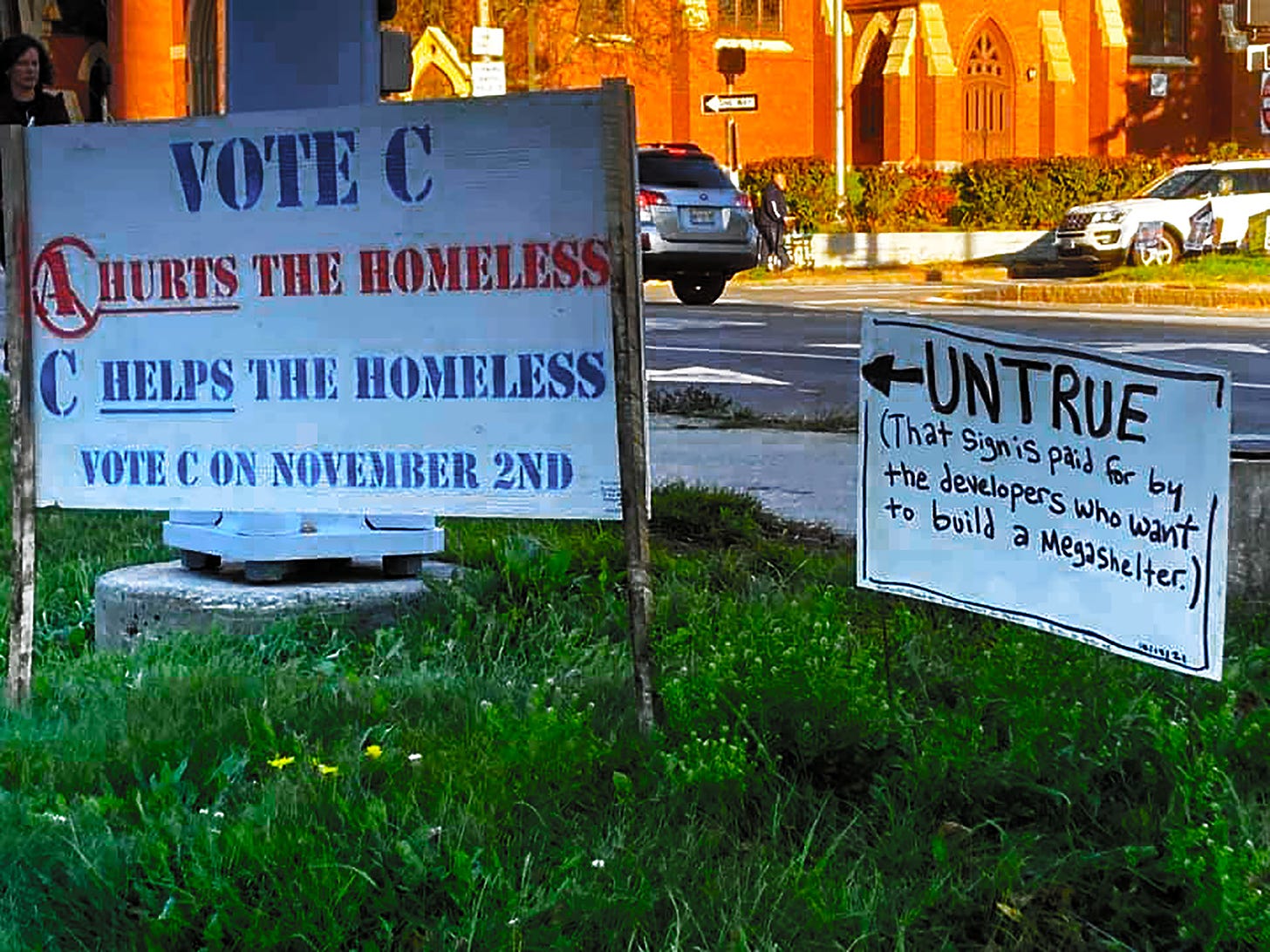 Large Sign says Vote C to support the Homeless, Small hand Made sign next to it says Umtrue! That sign is paid for by developers who want to build a megashelter!