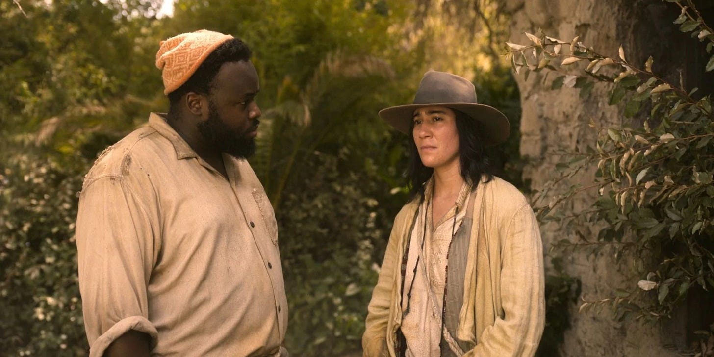 Oluwande, a black man with a beard, wearing an orange beanie looks at Jim, a nonbinary person with long hair, wearing a wide hat