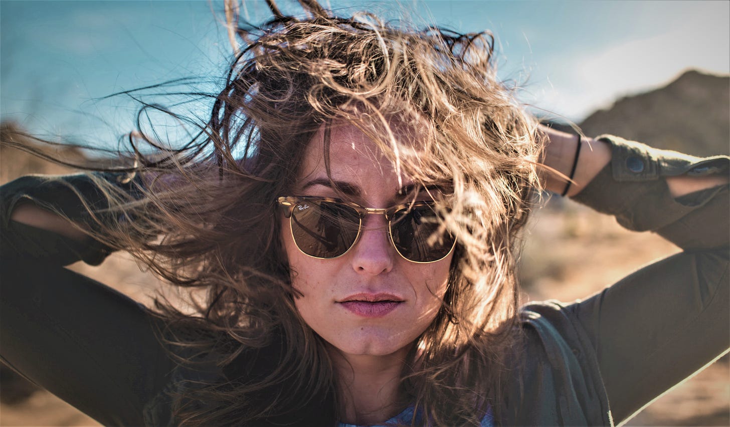 girl with brown hair wearing sunglasses and dark top with hands behind her head